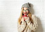 Smiling Hipster Girl in Knitted Sweater and Beanie Hat with Mug in Hands at White Brick Wall Background. Winter Warming Up Concept.