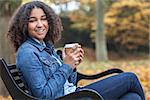 Beautiful happy mixed race African American girl teenager female young woman smiling and drinking takeaway coffee outside sitting on a park bench in autumn or fall