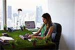 Concept of ecology and environment: Young business woman working in modern office with table covered of grass and plants. She types on tablet pc