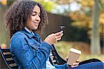 Beautiful happy mixed race African American girl teenager female young woman smiling drinking takeaway coffee and cell phone texting sitting on a park bench in autumn or fall