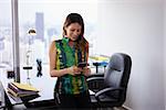 Young hispanic business woman leaning on table in modern office. She holds a mobile phone and is text messaging, smiling happy