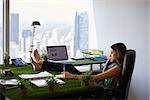 Concept of ecology and environment: Young business woman working in modern office with table covered of grass and plants. She writes on note pad with barefeet on desk