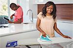 Woman ironing while boyfriend using tablet in the kitchen