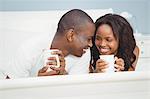 Ethnic couple holding mugs in bed in the bedroom