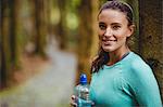 Fit brunette taking a break and holding bottle against a tree in the woods