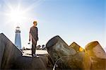 Young man holding skateboard, standing on breakwater near lighthouse, looking towards ocean