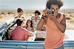 A group of friends by a pale blue convertible on the open road, on a flat plain surrounded by mountains, one holding a camera.