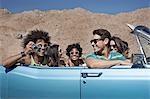 A group of friends in a pale blue convertible on the open road, driving across a dry flat plain surrounded by mountains.