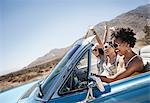Three young people in a pale blue convertible car, driving on the open road across a flat dry plain,
