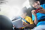 Two children travelling in the back seat of a car, one using a digital tablet and one a handheld game.