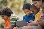 A row of children sitting outdoors in summer using tablets and handheld games.