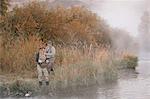A man and woman standing on a riverbank, fishing.