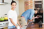 Young woman using laptop and her boyfriend baking in the kitchen at home