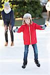 little boy learning ice skating and his mother watching and cheering up at outdoor skating rink, having winter vacation fun