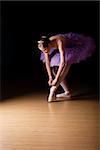 Young female ballerina wearing a lilac colored tut adjusting her shoes against black background