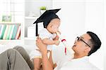Baby with graduation cap holding certificate with father. Parent and child early education concept. Asian family lifestyle at home.