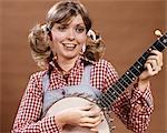 1970s YOUNG WOMAN IN CHECKED SHIRT AND OVERALLS WITH HAIR IN PONYTAILS PLAYING BANJO