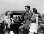 1930s TWO WOMEN IN NAUTICAL THEME CLOTHES AND MAN IN A SUIT TALKING STANDING BY THIRD WOMAN IN PARKED AUTOMOBILE AT SEASHORE