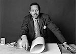1970s AFRICAN-AMERICAN BUSINESSMAN CONTRACTOR ARCHITECT BUILDER AT TABLE WITH BUILDING PLANS AND HARD HAT LOOKING AT CAMERA