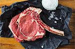 Rib-eye steak with a long bone on a piece of dark paper and pyramid salt