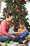 A mother and two children by a large Christmas tree unwrapping presents.