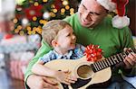 A father and son sitting by a Christmas tree, playing a guitar.