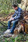 A man sitting on a tree stump plucking feathers from a game bird carcass.