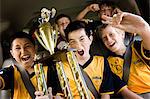 A group of boys in soccer team shirts holding a trophy and celebrating a win. In a team bus.