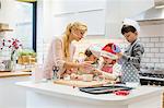 A woman and three children working together, making a gingerbread house, and icing the gingerbread.