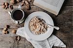 A table with a cup of coffee, bowl of muesli, nuts and an open book.
