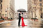 A woman in a long red evening dress with fishtail skirt and a fur stole, and a man in a suit, kissing under an umbrella in the city.