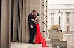 A woman in a long red evening dress with fishtail skirt and a fur stole, and a man in a suit kissing on the steps of a building.