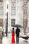 A woman in a long red evening dress with fishtail skirt and a fur stole, and a man in a suit, walking through snow in the city.
