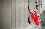 A woman in a long red evening dress with fishtail skirt and a fur stole, and a man in a suit hand in hand on the steps of a building.