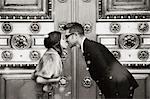 A couple, man and woman kissing in front of the large decorated doors of a city building.