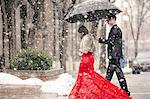 A woman in a long red evening dress with fishtail skirt wearing a fur stole, and a man in a suit, walking through snow in the city.