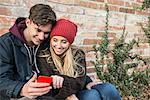 Young couple smiling and using smart phone against brick wall, Munich, Bavaria, Germany