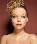 beauty shoot of very pretty woman with glossy golden creative make-up and accessory in the hairdo. She is covering her nude breast and looking in camera