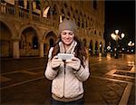 Evening promenade in Venice take you into another world. Smiling young woman tourist writing sms while standing on St. Mark's Square near Dogi Palace in the evening