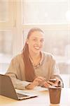 Toned Portrait of happy freelance woman looking at camera while sitting at table in front of laptop computer. Smiling lady working in cafe or office interior.