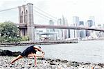 Young man practicing yoga on riverside in front of Brooklyn Bridge, New York, USA