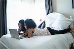 Lesbian couple lying on bed, using laptop