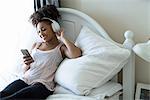 Woman relaxing on bed, wearing headphones, looking at smartphone