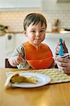 Baby boy sitting at table being fed yogurt by mother