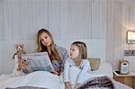 Mother reading newspaper beside daughter in bed