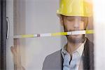 Young businessman wearing yellow hard hat measuring window in new office