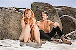 Couple sitting on beach, against rocks, laugh