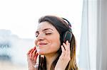 Beautiful young woman listening to headphone music in front of  apartment window