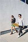 Young couple walking together outdoors, woman using smartphone, man carrying skateboard