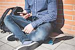 Neck down view of young male urban skateboarder sitting on sidewalk reading smartphone text